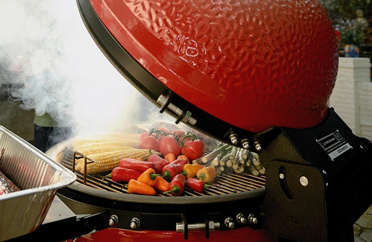 Smoke billows out of an open Kamado Joe grill filled with a variety of vegetables. The view shows the grill from the side and rear to show the Air Lift hinge. 
