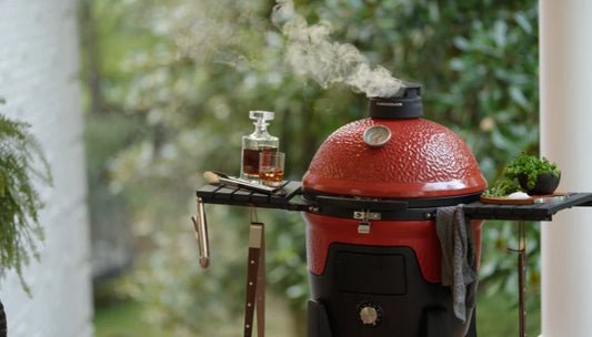 Smoke rises from the top vent of a Pellet Joe grill. The left shelf holds a filled crystal decanter with matching glass, tongs, and a grill gripper. Fresh green herbs and a pile of salt sit on a wooden board on the right shelf.