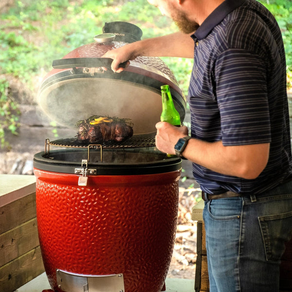 Smoke escapes from a Classic Joe III standalone grill installed in an outdoor kitchen as a man holding a drink opens the lid to check on a roast topped with fresh herbs and lemon slices.