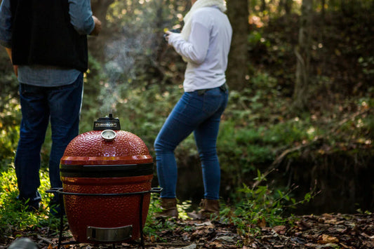 2 people dressed for chilly fall weather stand next to a Joe Jr grill outside  in a wooded area