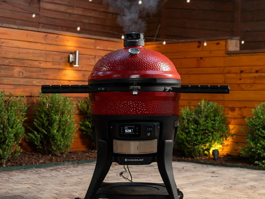 A Big Joe Konnected Joe Digital Charcoal Grill and Smoker outside on a brick patio in front of bushes and 2 stained wood fences. Smoke escapes from the Kontrol Tower top vent. 