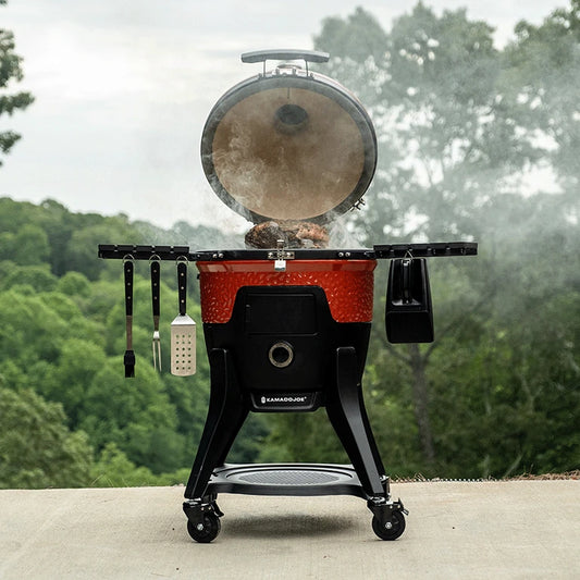 A rack of pork chops cooks on the grate of a Pellet Joe grill with smoke curling up around it. Cooking utensils hang from hooks on the left shelf and a pellet scoop hangs off the right shelf.