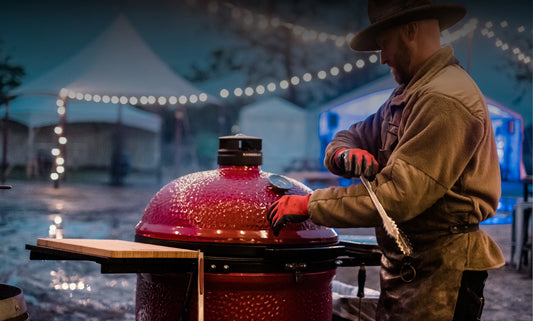 Chef Eric Gephart ears a heavy coat, hat, and gloves while grilling on a cold and rainy day. A string of lights put up in front of several tents can be seen reflected on wet pavement.