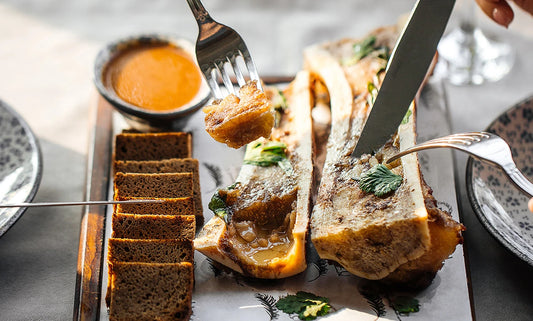 2 forks and a knife are used to remove smoked bone marrow from 2 bones split in half lengthwise. A row of bread squares and a bowl of dipping sauce are also on the serving platter.