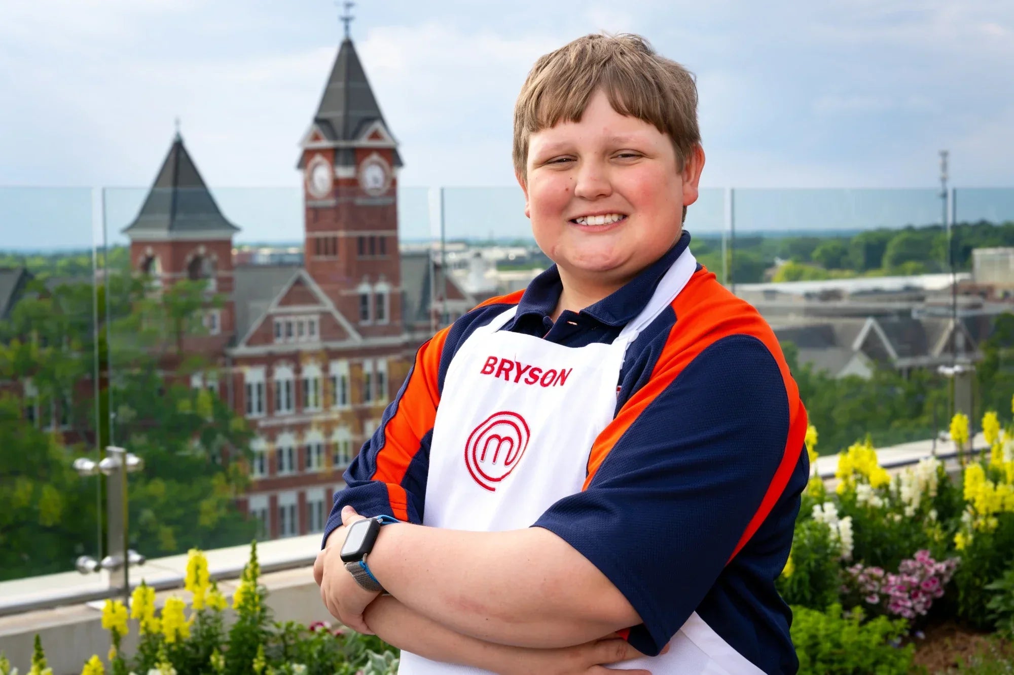 Bryce McGlynn wearing a 'Bryson' apron with a logo, standing in a rooftop garden overlooking Auburn University's Samford Hall