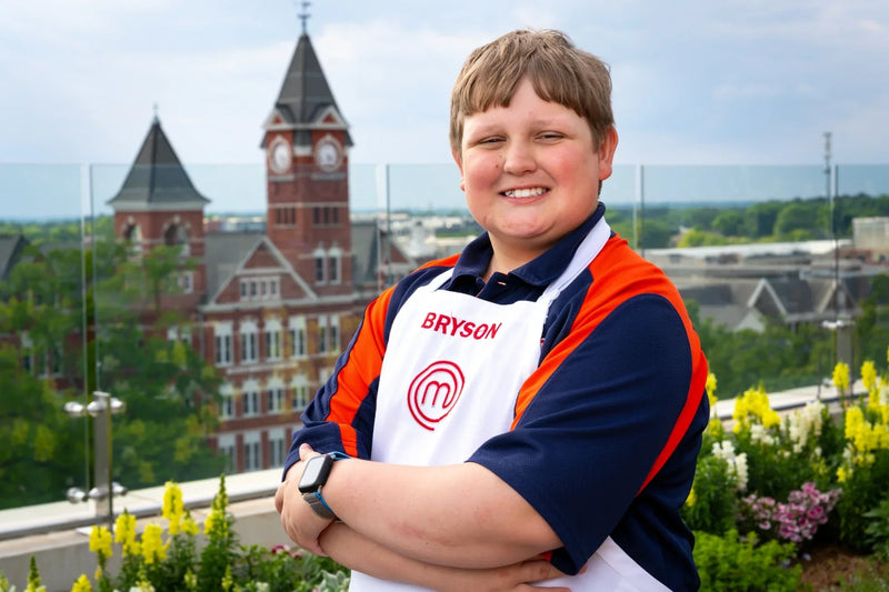Bryce McGlynn wearing a 'Bryson' apron with a logo, standing in a rooftop garden overlooking Auburn University's Samford Hall