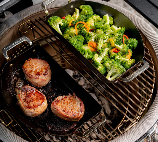 The Karbon Steel half-moon pan set in use on a 2-tier Divide & Conquer rack. The solid pan is on a lower, hotter rack and holds 3 searing steaks. The perforated pan sits on the higher, cooler rack full of broccoli florets and other vegetables.