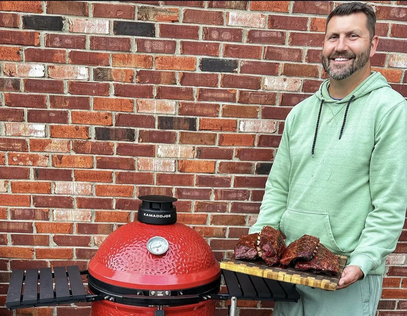 Ambassador Jace Green holding a tray of cooked ribs next to a Kamado Joe grill against a brick wall.
