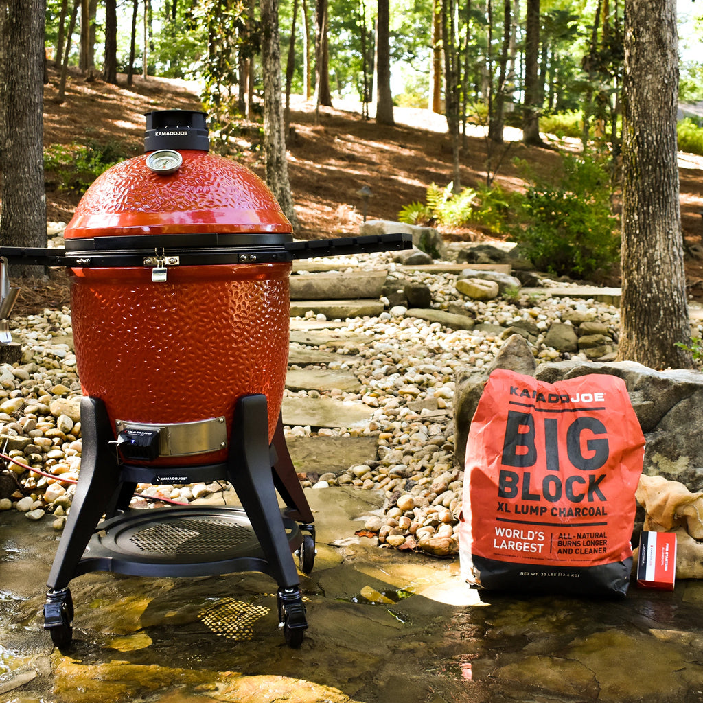 A closed Kamado Joe grill site on a stone-paved patio next to a bag of Big Block XL Lump Charcoal and a box of Kamado Joe Fire Starters. A stone path behind the grill leads into a wooded hillside.