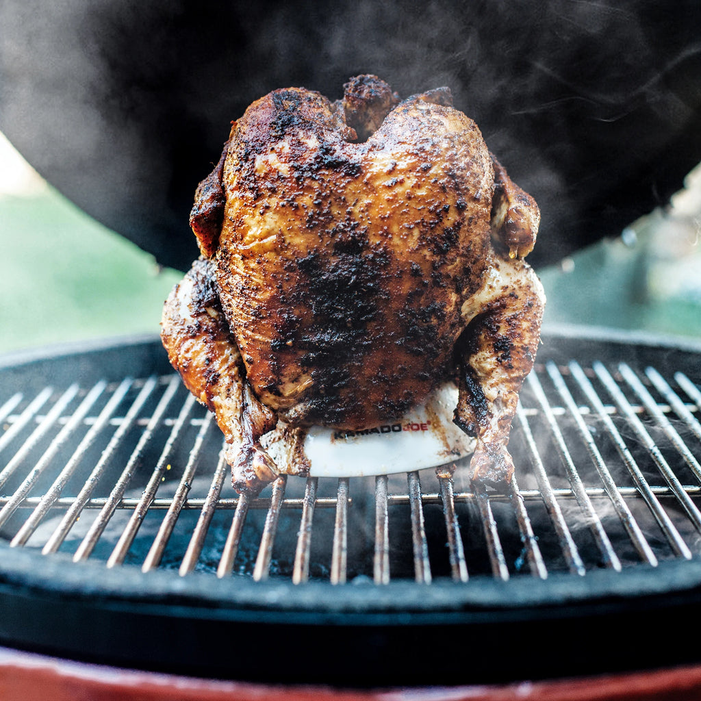 A whole chicken sitting upright on a Kamado Joe chicken stand on the grate of a Kamado Joe grill. 