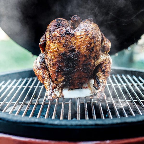 A whole chicken sitting upright on a Kamado Joe chicken stand on the grate of a Kamado Joe grill. 