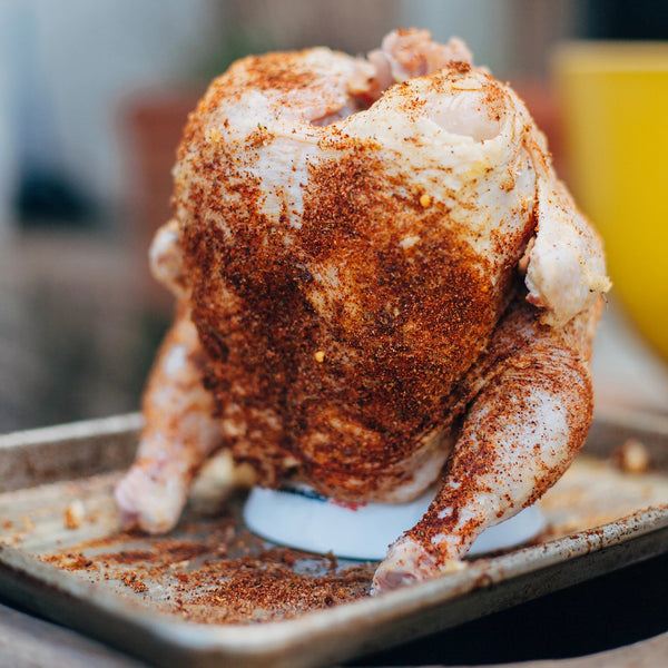 A seasoned whole chicken sits ready to cook with legs down on a chicken stand in a metal tray