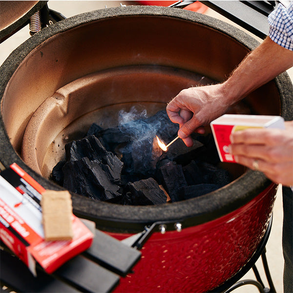 A man reaches into an open Kamado Joe grill as he holds a lit match near the pile of XL Big Block  charcoal in the bottom of the grill. 