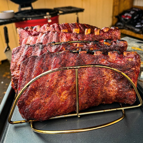 4 cooked racks of ribs stand in a Kamado Joe rib rack set in a metal tray with an open Kamado Joe grill in the background.