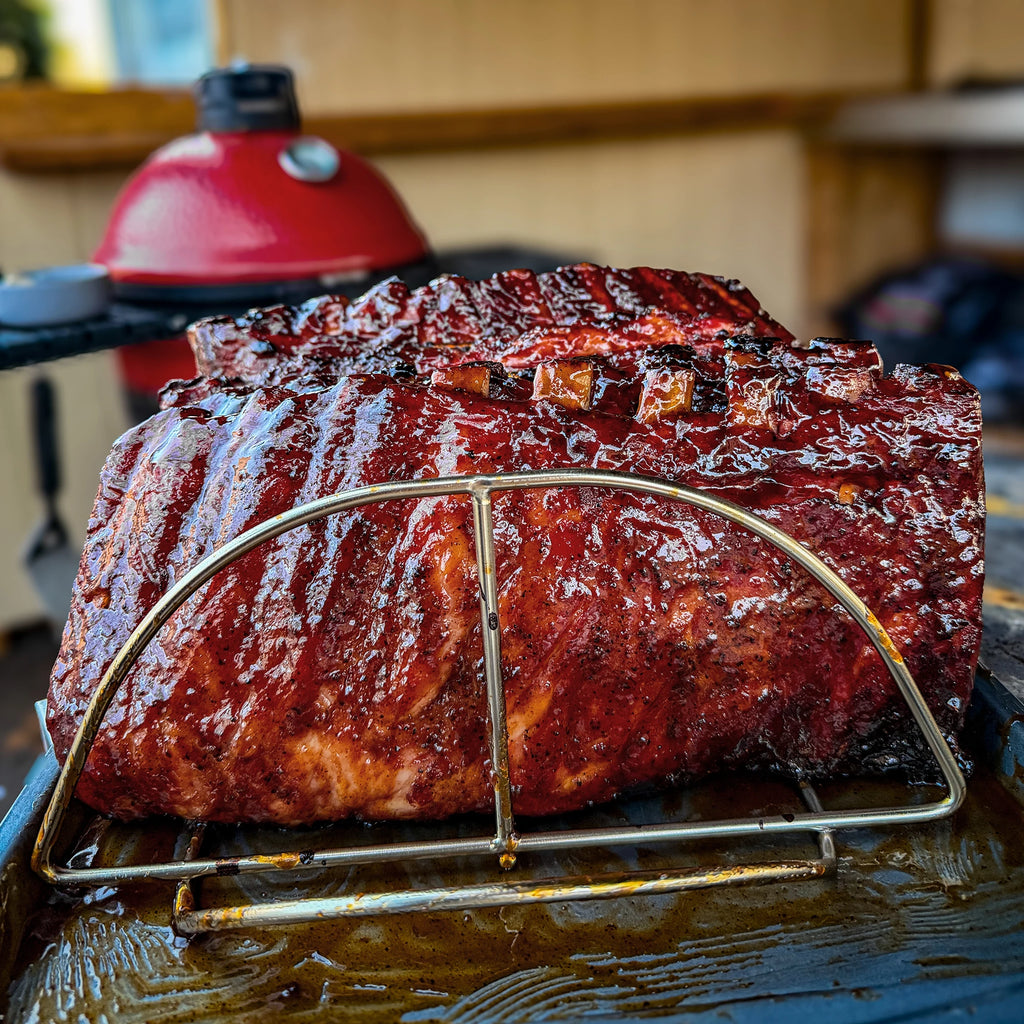 2 cooked racks and sauced racks of ribs stand in a Kamado Joe rib rack set in a metal tray.