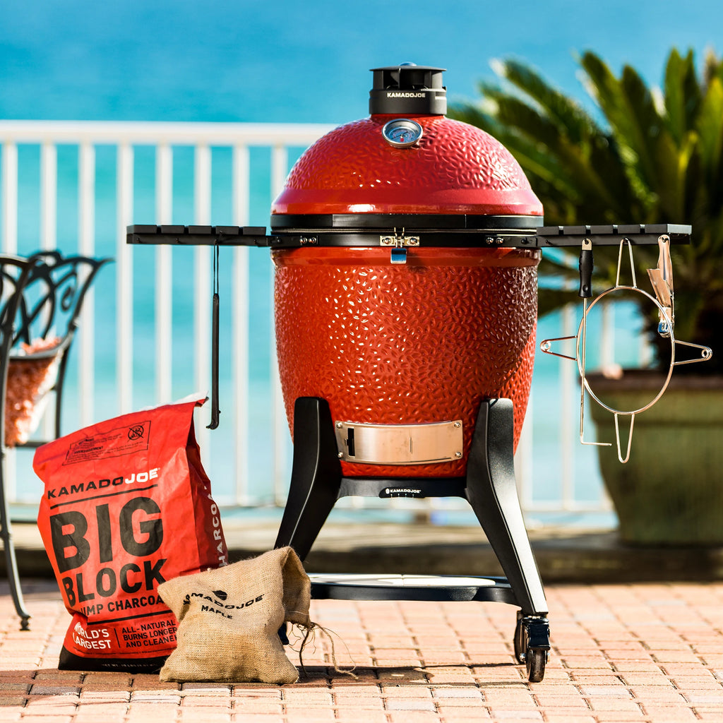 A Kamado Joe Grill sits on an outside deck overlooking the ocean. A bag of Big Block lump charcoal and a bag of hickory wood chunks sit on the patio next to the grill.