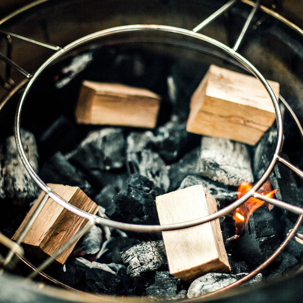 4 wood chunks sit on top of a pile of lump charcoal in the firebox of a Kamado Joe grill. There is a small flame burning on one block of charcoal. An accessory rack sits above the charcoal and wood chunks.