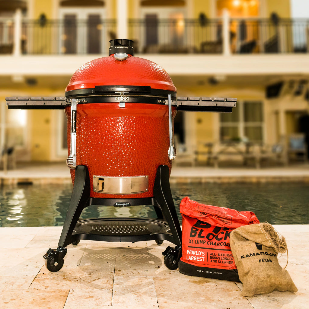 A Kamado Joe grill on a paved patio at the side of a pool. A partially used bag of Big Block lump charcoal with the top rolled down and an open bag of wood chunks sit on the ground next to it.