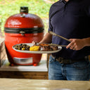 A man holds a plate of grilled foot in one hand and tongs in the other.  He is standing in an outdoor kitchen with a Big Joe III Standalone grill installed in a low area of the counter behind him.