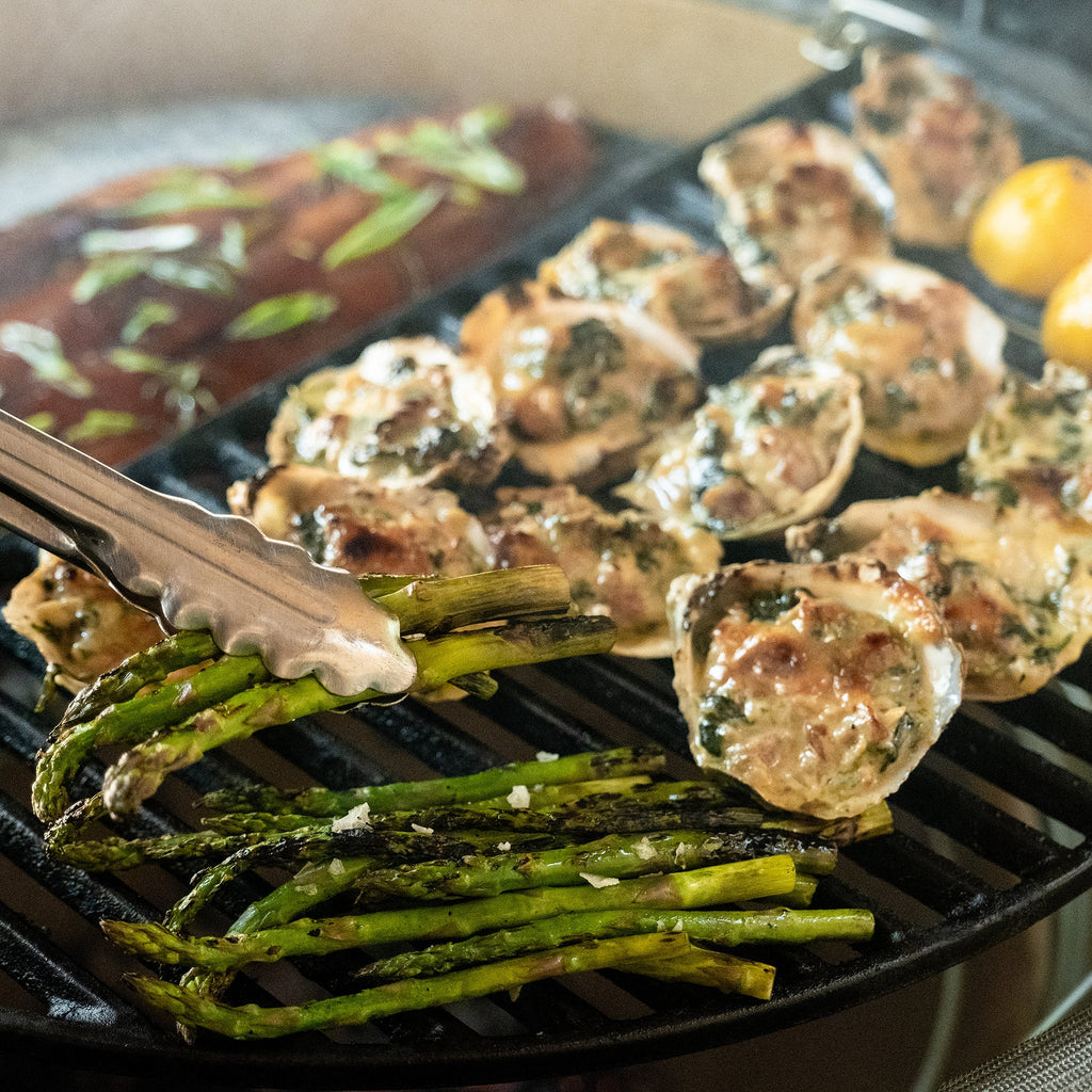 Someone uses tongs to turn asparagus cooking on a grill along with oysters in the half shell and halved lemons. In the background a large fish filet topped with fresh herbs cooks on a half-moon soapstone surface on a lower tier of the Divide & Conquer system.