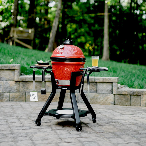 A Joe Jr sits in the cart with shelves on a paved outdoor patio in front of a stone block retaining wall and s sloped lawn. The cart shelves hold tools, a dish with onion slices and herbs, and a nearly full drinking glass.