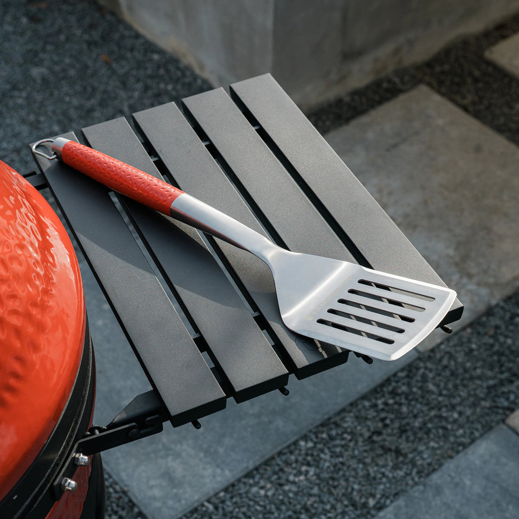 A stainless steel Kamado Joe spatula rests on the side shelf of a Kamado Joe grill. The spatula has a red dimpled handle.