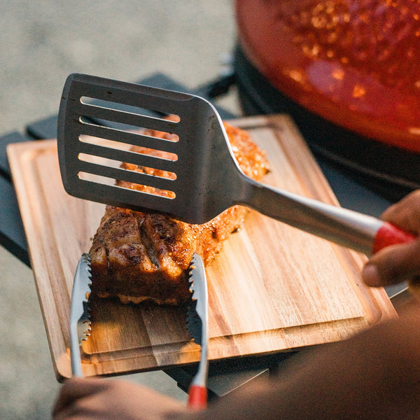 Person using a Kamado Joe spatula and tongs to handle a cooked salmon filet on a wooden cutting board on the side shelf of a Kamado Joe grill
