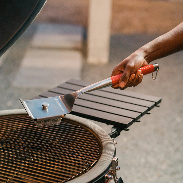 A person uses the bristles of a Kamado Joe grill brush to clean the grate of a Kamado Joe grill.