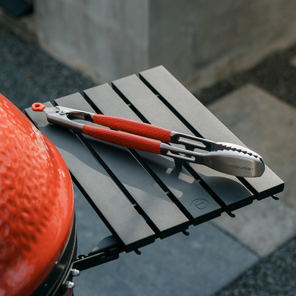 Steel tongs with Kamado Joe red handles sitting on the side shelf of a Kamado Joe grill. The tongs are positioned to show the grooved teeth.