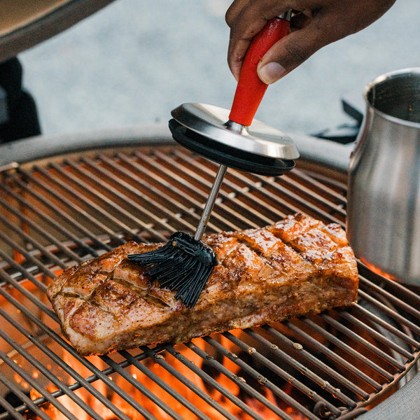 Person using a Kamado Joe basting brush to apply sauce to salmon on a grill. The basting pot sits on the grate next to the salmon.