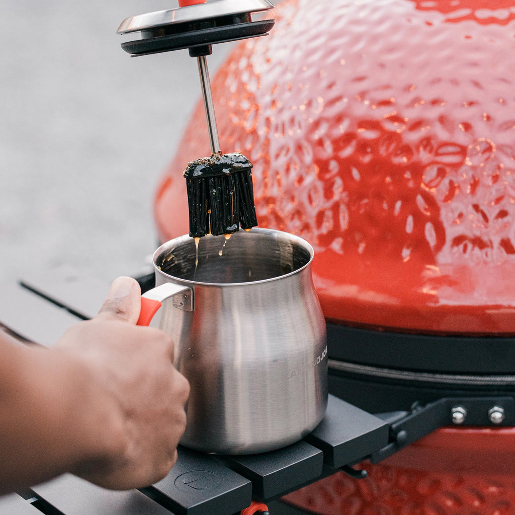 Person holds the basting brush above the basting pot sitting on the side shelf of a Kamado Joe grill. The person holds the pot by its handle to keep it steady.