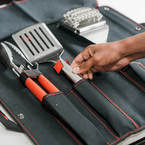 Someone places a Kamado Joe spatula in the sleeve of the canvas tool kit case. A pair of tongs and a grill brush are already in other pockets of the case. 