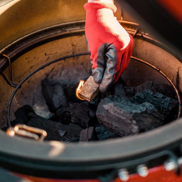 A person wearing a heat-resistant grilling clove lowers a lit Kamado Joe Fire Starter block into a pile of big block lump charcoal inside a Kamado Joe grill