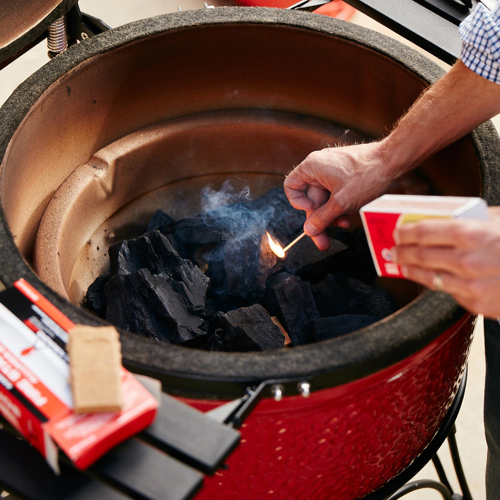 A man lowers a lit match into a pile of big block lump charcoal inside a Kamado Joe grill