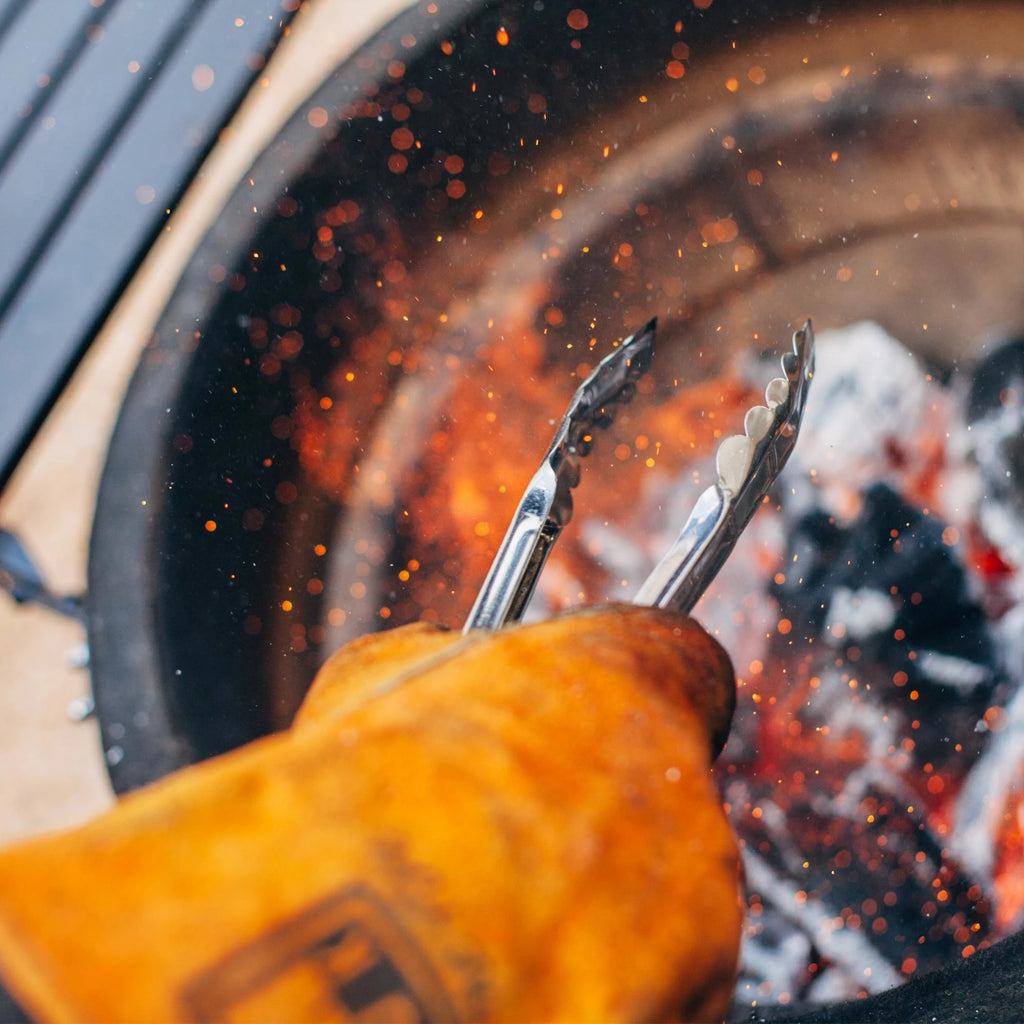 A person wearing a leather grilling glove uses metal tongs to spread burning charcoal in the firebox as sparks rise up from the coals.