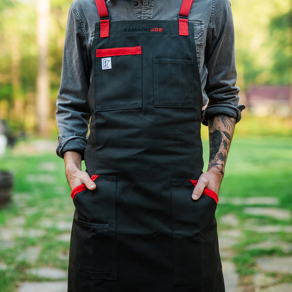 A man wearing a Kamado Joe apron stands outside. His torso and upper legs are covered by the apron. He stands with his hands in the 2 side pockets. The background is blurred.