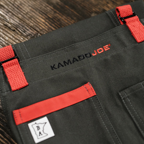 A folded apron rests on a dark wooden surface, shown as a close-up highlighting the embroidered Kamado Joe name and the orange accent color used for the straps and as a highlight across the top of one pocket.