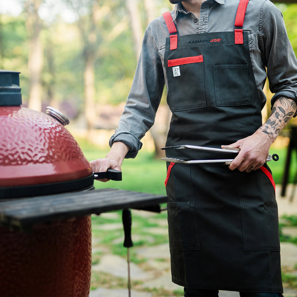 A man wearing a Kamado Joe apron hold the handle of a Kamado Joe grill with his right hand and a set of metal tongs with his left. His torso and upper legs are visible showing complete coverage by the apron.