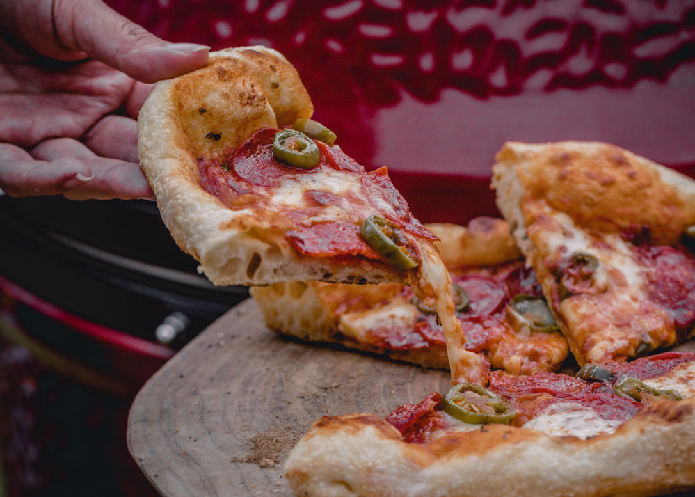 Someone pulls a slice out of a pizza topped with cheese, pepperoni, and jalapeno slices. A red Kamado Joe grill is in the background.