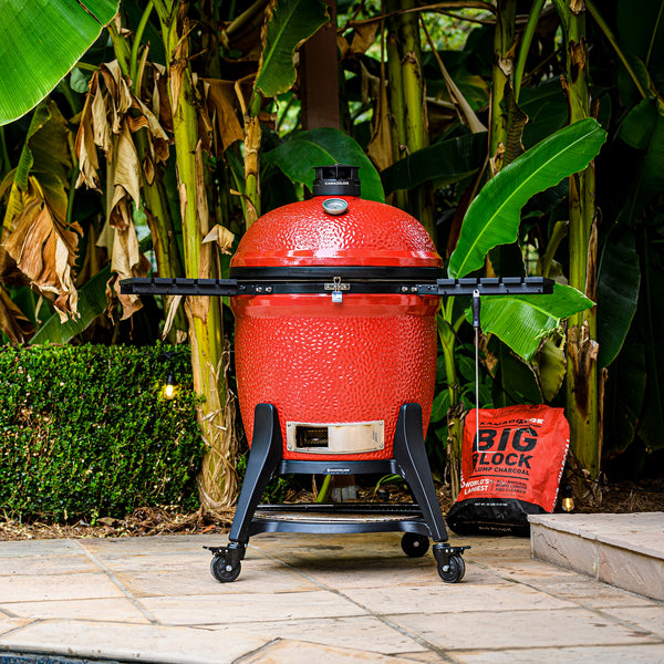 A Big Joe III grill sits on a paved patio in front of tropical greenery and a bag of Kamado Joe Big Block XL Lump Charcoal
