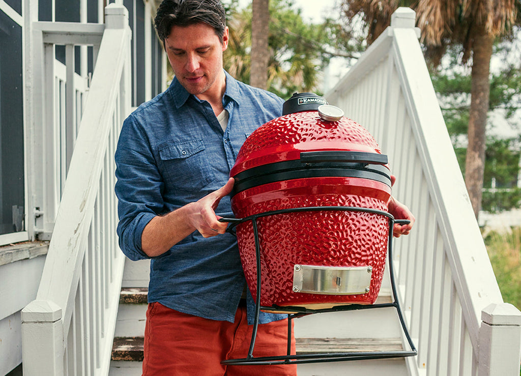 A man in a blue shirt and orange shorts uses the side handles of the grill stand to carry a Joe Jr grill down stairs outside of a home.